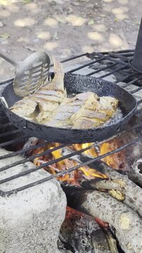 Pescado entero fresco fri&eacute;ndose en una sart&eacute;n de hierro fundido sobre una hoguera de le&ntilde;a, con aceite hirviendo y brasas brillantes, mostrando la cocina r&uacute;stica al aire libre, carb&oacute;n encendido