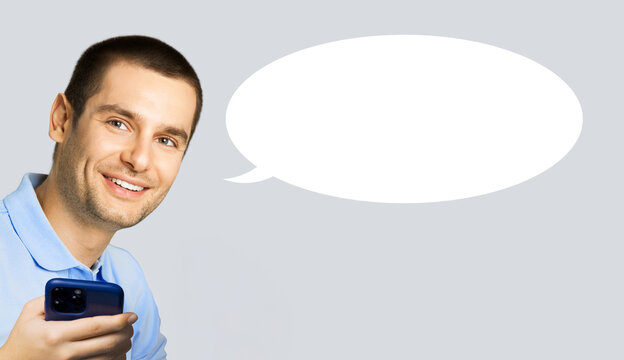 Portrait image - happy smile young man wear blue t-shirt cloth, holding hand cell phone mobile smartphone cellphone, isolated grey gray background. White empty mockup speech bubble with free space