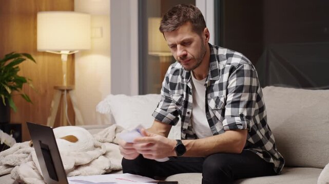 Man sitting on couch at home holding paper bills and financial documents near a laptop. Concept of financial pressure from increasing household expenses and difficulty managing payments.