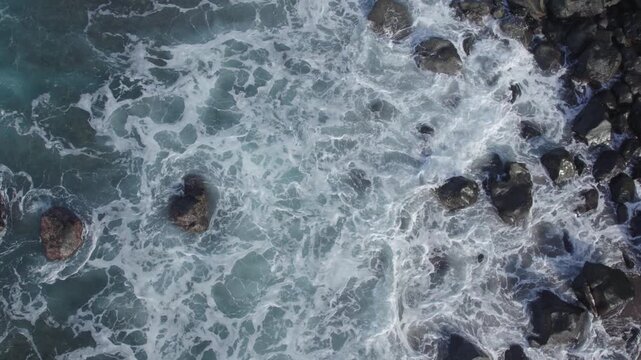 Cinematic slow motion footage of powerful ocean waves hitting dark volcanic rocks, creating beautiful white spray and foam on the coast of Flores.
