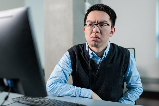 Young Asian man looks at computer with a skeptical expression in the office, ideal for AI anxiety, job insecurity, or technology distrust concepts.