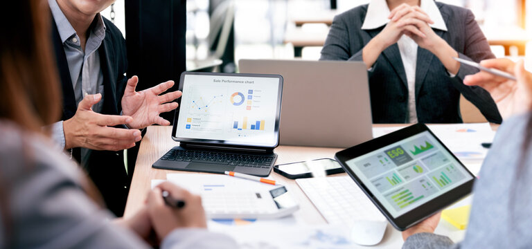 Business team analyzing financial sales performance charts and ESG reports on digital tablets and laptop during a collaborative meeting in the office.
