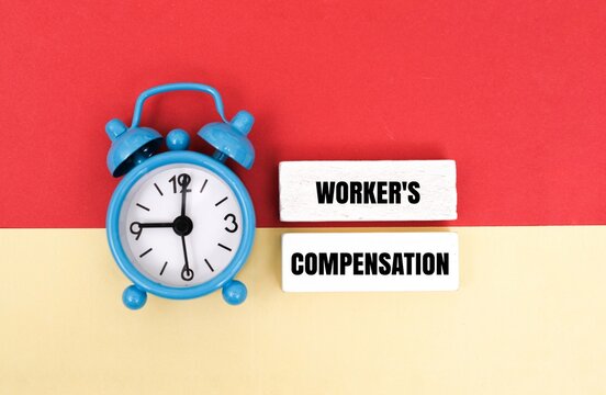 A blue alarm clock next to wooden blocks with text WORKER'S COMPENSATION on a colorful background. Concept for workplace injury insurance, legal deadlines and employee rights