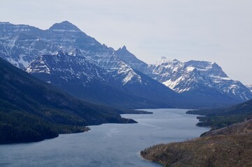 Scenic view of Waterton Lakes National Park, showing rocky mountains with snow patches and a calm lake. The natural landscape captures tranquil water, trees, and a serene, scenic environment.