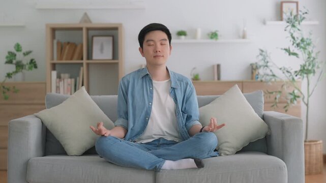 Happy young Asian man practicing yoga and meditation at home sitting on floor in living room in lotus position and relaxing with closed eyes. Mindful meditation and wellbeing concept