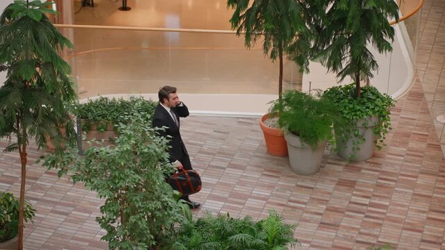 High angle view of Caucasian businessman in suit talking on mobile phone while walking with brown leather briefcase amid potted plants palms and modern curved glass atrium interior office building