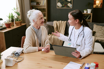Obraz premium Senior Caucasian woman sitting at table interacting with young adult Caucasian female doctor holding digital tablet, discussing health care in home environment