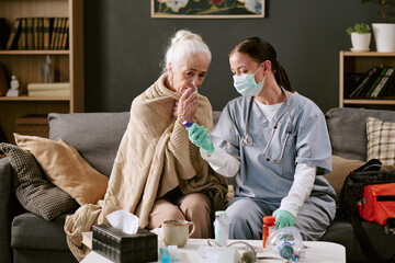 Senior Caucasian woman sitting on sofa wrapped in blanket being assisted by young adult Caucasian...
