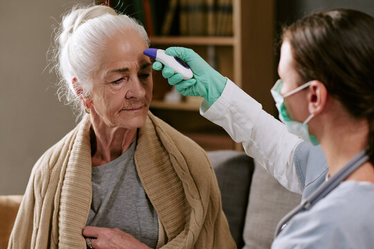 Senior Caucasian woman sitting while female healthcare worker measuring forehead temperature with digital thermometer, medical examination taking place in clinical setting