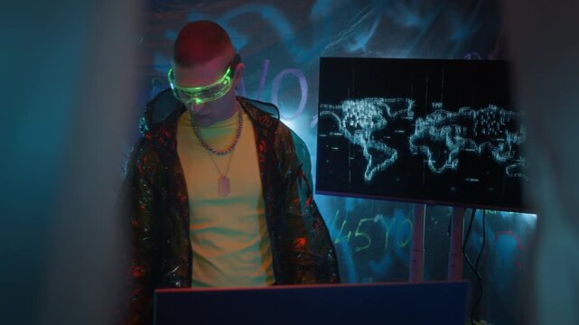 Tracking shot of young man wearing black transparent coat and neon visor using computer system while hacking in dark workspace