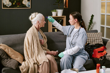 Senior Caucasian woman sitting on sofa being examined by young adult Caucasian female nurse wearing medical gloves and scrubs, nurse measuring temperature with digital thermometer
