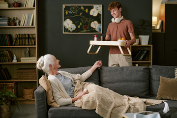 Senior Caucasian woman reclining on sofa under blanket interacting with smiling Caucasian teenage...