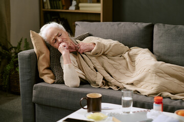 Senior Caucasian woman lying on couch under blanket, appearing unwell and resting with hand near...