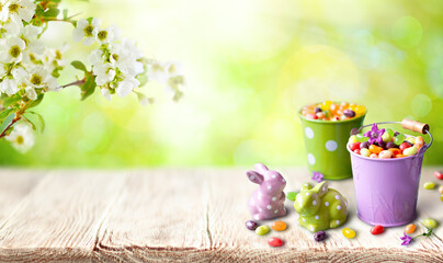 A cheerful spring setup featuring candy-filled pastel buckets, ceramic bunny figurines with polka dots, and blooming white blossoms in the background.