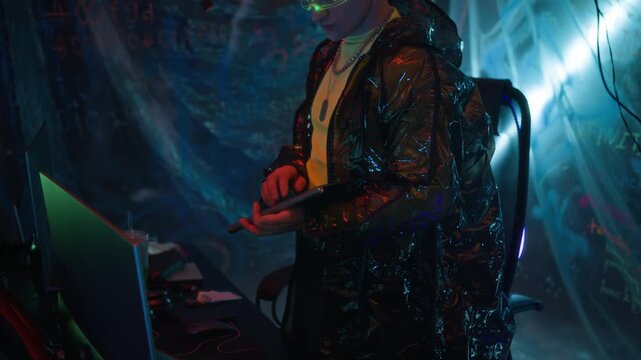 Tilt up shot of futuristic young male hacker wearing visor and reflective jacket using keyboard while stealing data on computer system in dark neon underground room