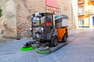Street cleaning vehicle with rotating brushes operated by municipal worker cleaning urban road surface, maintenance machine moving through historic city street environment