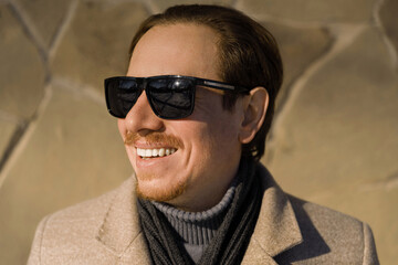 Fototapeta premium Close-up portrait of a young caucasian redhead man with sunglasses smiling wearing a coat on a winter day while walking down the street. Lifestyle.