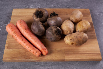 The ingredients for cooking carrots, potatoes and beets are on a wooden cutting board.