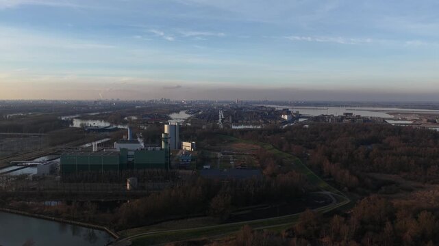 High angle aerial view showing a large waste to energy plant processing refuse derived fuel, located near a river and an industrial port complex with the city skyline in the distant background