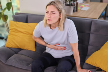 Obraz premium A woman sits on a gray couch with yellow cushions, holding her stomach with a look of distress. Sunlight fills the cozy living room, creating a warm atmosphere.