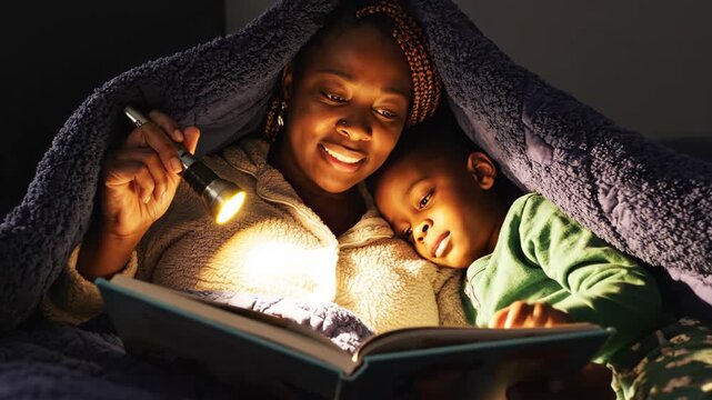 African American woman reading bedtime story to young son under blanket with flashlight. Cozy family bonding moment illuminated by soft light in intimate nighttime setting