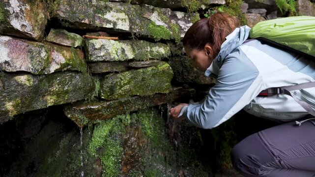 A young woman in her 30s squats in front of a rock wall from which fresh water springs in the mountains, drinking with her hands, dressed in mountain clothing and carrying a backpack.