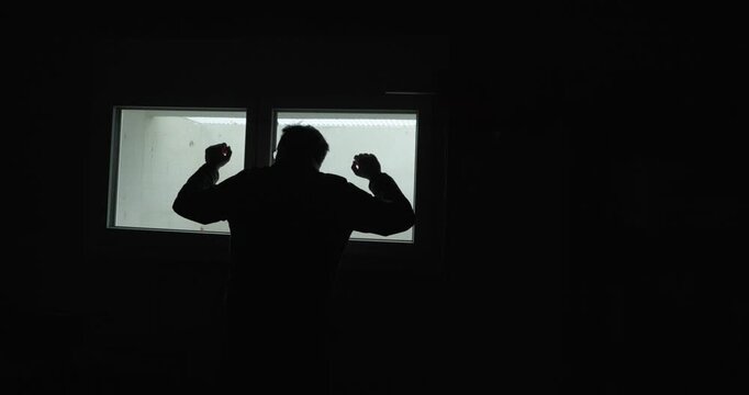 Moody silhouette of a man standing in front of a bright window, raising his fists in frustration inside a dark room. Ideal for concepts like anger, stress, conflict, isolation, mental health, and emot