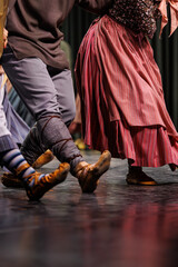 A dynamic close-up of traditional dancers in motion, showcasing striped skirts, patterned socks, wrapped leggings, and moccasin-style shoes as they perform a synchronized folk dance on stage