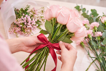 Florist's hands arrange a bouquet of roses and tie a red ribbon, with other flowers lying around. The concept of preparing a bouquet for a holiday or celebration.