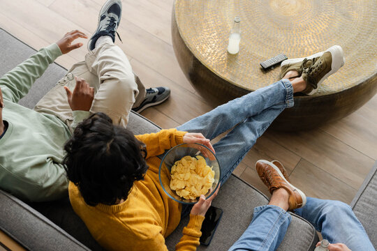 Diverse male friends sitting on gray couch at home wearing mustard knit holding chips bottle remote
