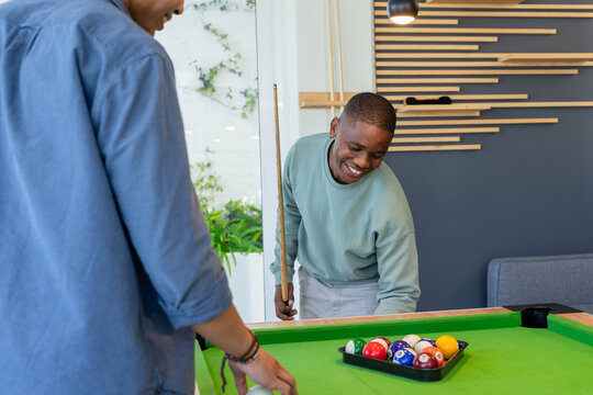 Diverse men in green sweatshirt, blue-shirt placing rack on pool table in game room, holding cues