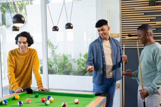 Diverse male friends leaning, holding cues, preparing shot around green pool table in rec room