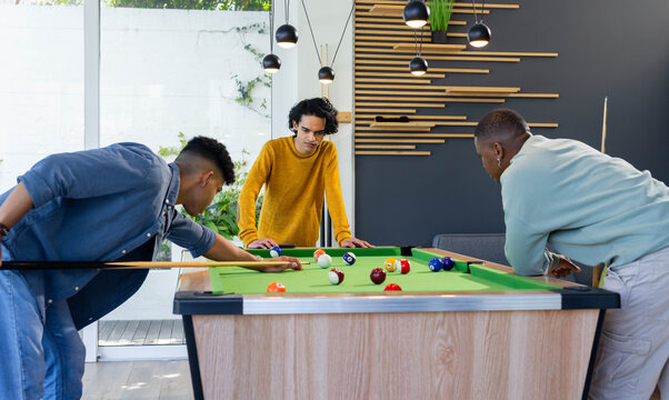Diverse male friends lining up shots around pool table with cues and billiard balls in lounge