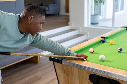 African American man leaning on green-felt pool table at home, lining up shot with cue stick