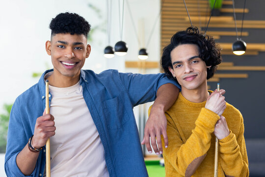 Diverse male friends standing by green pool table wearing denim and yellow sweater, holding cues
