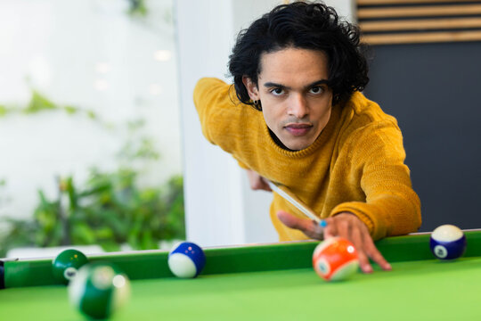 Man leaning over green baize pool table in lounge aiming shot with cue, wearing mustard sweater