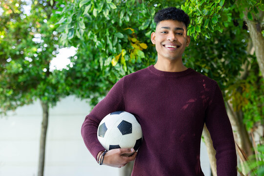 Adult male standing smiling holding soccer ball in garden wearing maroon sweater and bracelets