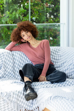 African American woman in 20s sitting on sofa with houndstooth throw, wearing rose top and boots