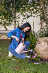 Asian woman pouring water to flowering plants beside rock with trowel on grass in garden