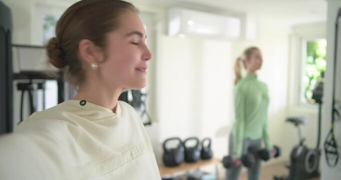 Female vlogger greeting viewer while friend curling dumbbells in home gym for workout tips