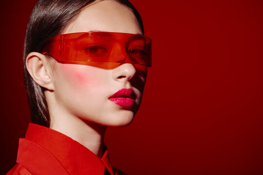 Close-up portrait of a woman wearing translucent red visor glasses and vibrant lipstick. Red lighting emphasizes cheeks and skin texture. Concept for beauty, fashion, and creative style.