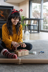 Naklejka premium Woman wearing festive headband wrapping presents on rug in living room near fireplace using ribbon