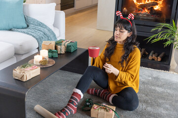 Naklejka premium Woman wearing mustard sweater and candy cane headband wrapping gifts at home sipping, mug