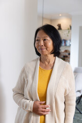 Naklejka premium Senior Asian woman standing inside living room next to column, wearing beige cardigan, mustard top