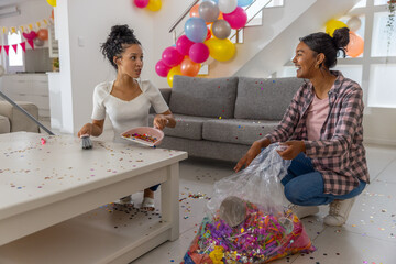 Diverse mom and teenage daughter sweeping confetti into pink dustpan and bag in living room
