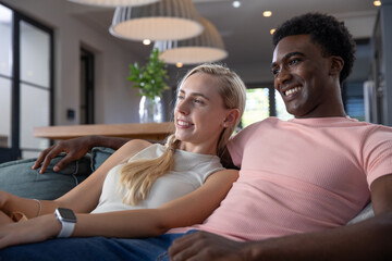 Diverse couple sitting on sofa in living room, smiling and wearing smartwatch next to glass vase
