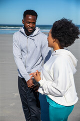 African american couple in athletic wear talking on sandy beach near waterline with smartphone