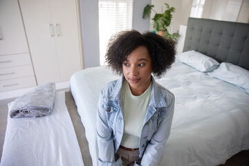 African American woman in her 30s sitting on bed edge in bright bedroom wearing denim jacket