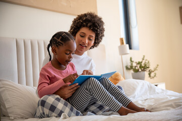 African american mother with daughter sitting on bed reading blue children book in bedroom