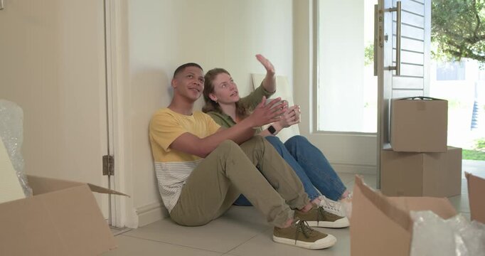 Diverse couple sitting on entryway among moving boxes bubblewrap with mugs pointing planning layout